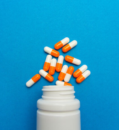 A white plastic cylindrical medical pill bottle with the grooves of a screw top at the neck of the vessel lies on its side. Between 10 and 20 oblong orange and white pill capsules are arranged as if they've spilled out of the open bottle. Bottle and pills alike are on a light blue background which takes up the entire frame of the photo