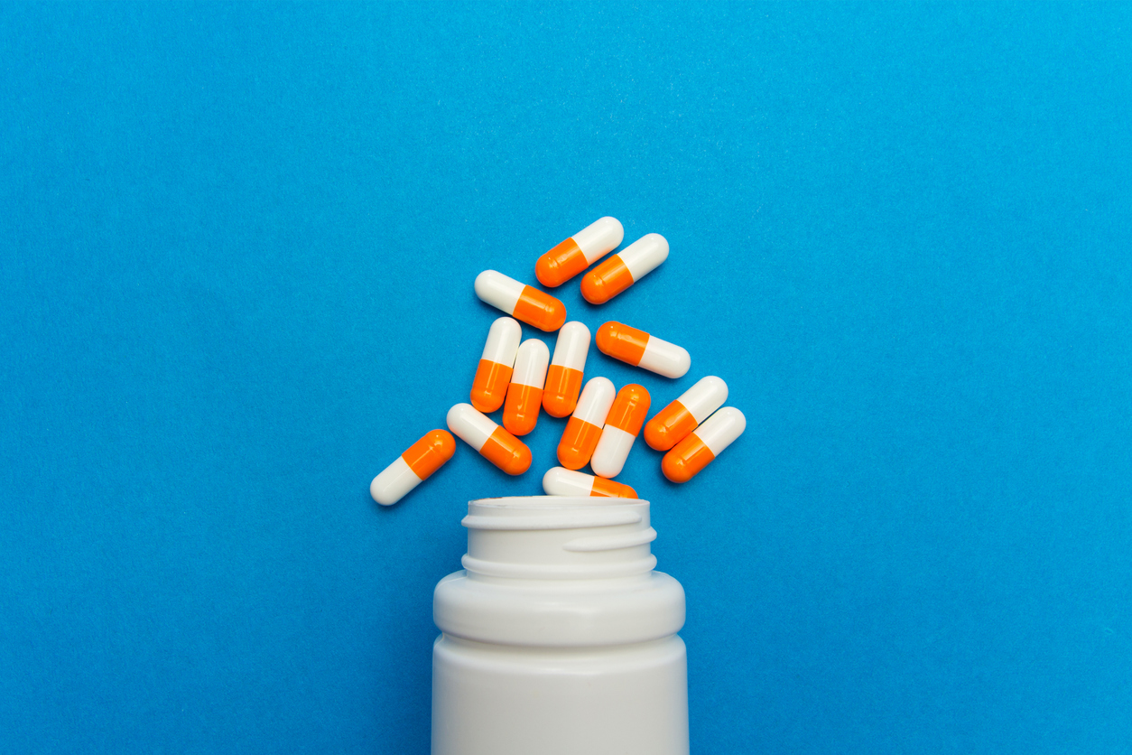 A white plastic cylindrical medical pill bottle with the grooves of a screw top at the neck of the vessel lies on its side. Between 10 and 20 oblong orange and white pill capsules are arranged as if they've spilled out of the open bottle. Bottle and pills alike are on a light blue background which takes up the entire frame of the photo