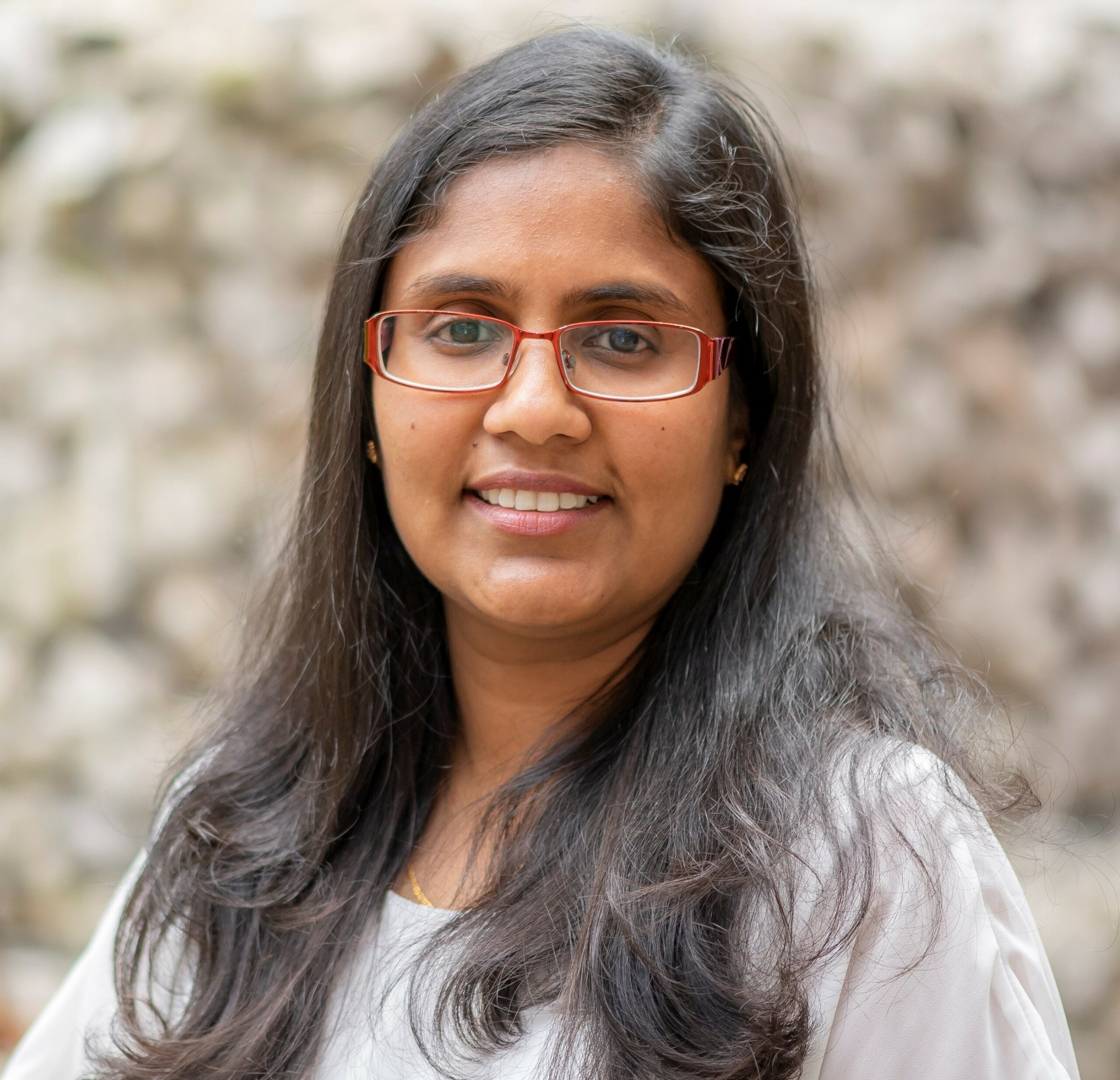 Close cropped head and shoulders profile picture of Dr. Monalie Bandulasena. She is a south asian woman descended woman in her late 30s with shoulder length dark hair and red medium width framed glasses wearing a light grey blouse