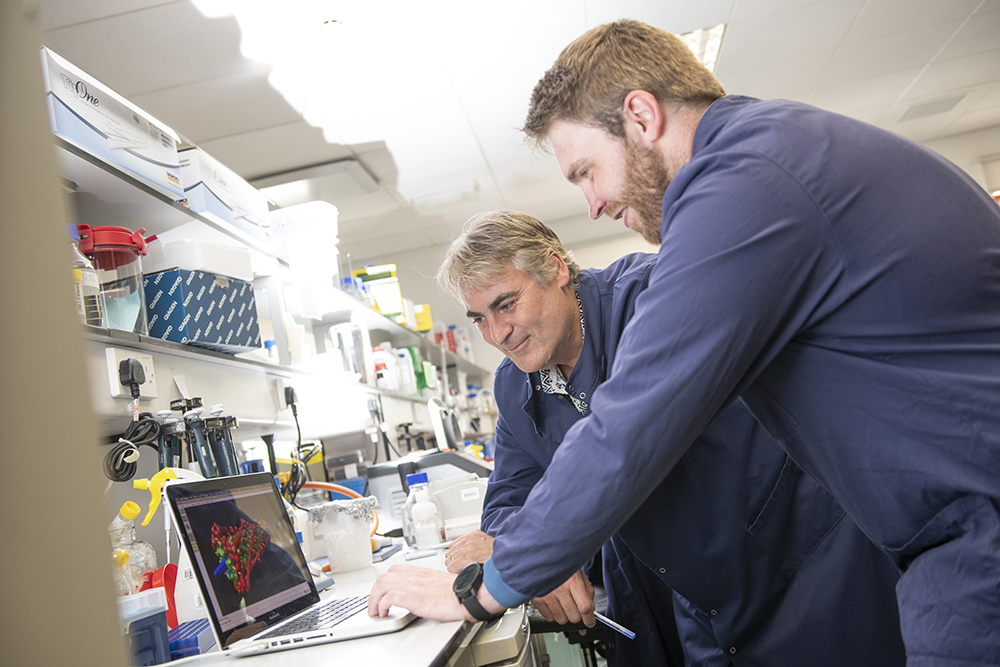 Picture of two of the NanoSyrinx founders. Both are white men, one in his fifties the other in his thirties. They are in a laboratory setting, wearing blue overalls and leaning on a work bench to look at a laptop screen which displays a 3d graphic